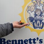 Lego Ben in his wheelchair is being held next to the Future Home of Bennett's Village sign. Lego Ben has brown, wavy hair, a huge smile, and a red shirt.