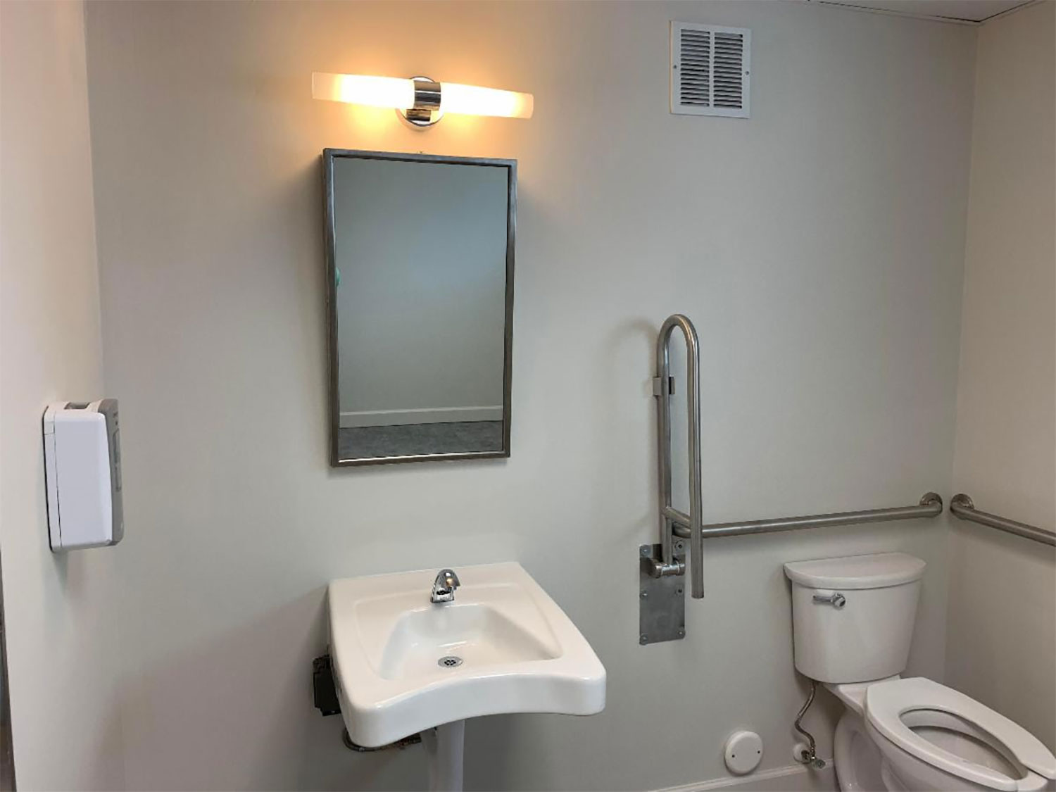 A bathroom mirror, sink, and toilet with grab bars around it