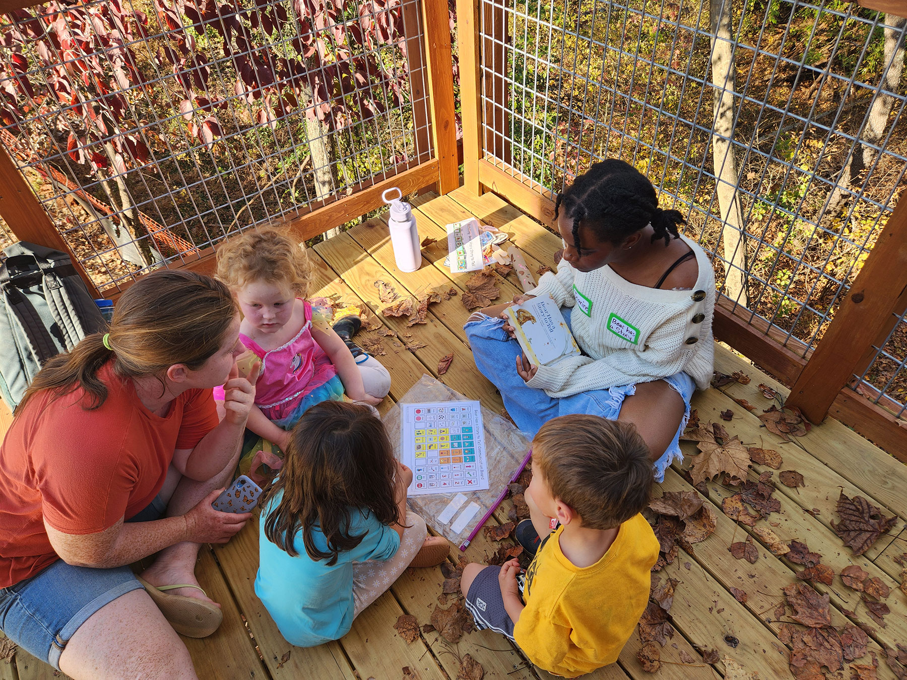 A student from UVA School of Education and Human Development sits on the deck of the accessible treehouse at Bennett's Village, reading to a group of children.