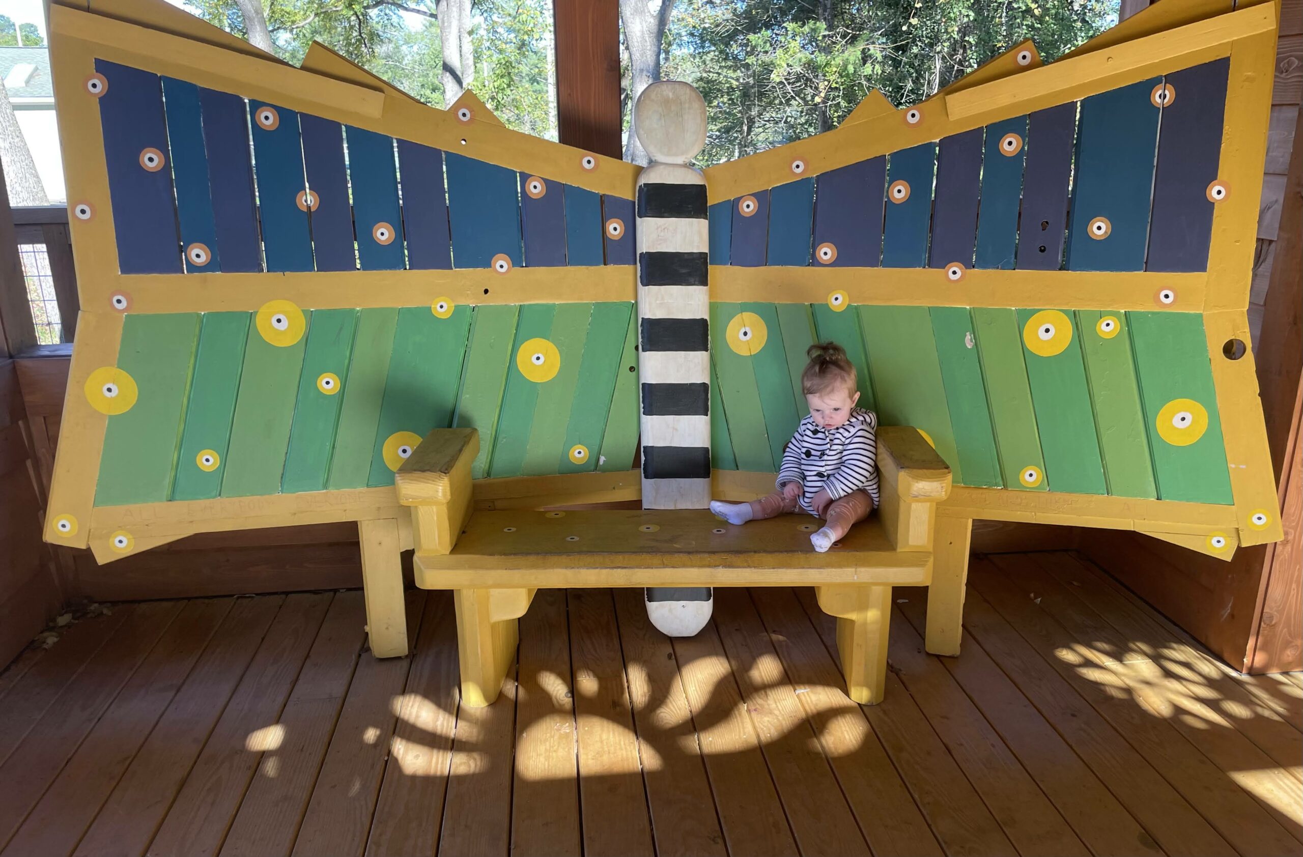 A child sits on the blue, green, and yellow butterfly bench.  Shadows from the woodwork project a sun on the floor of the treehouse.